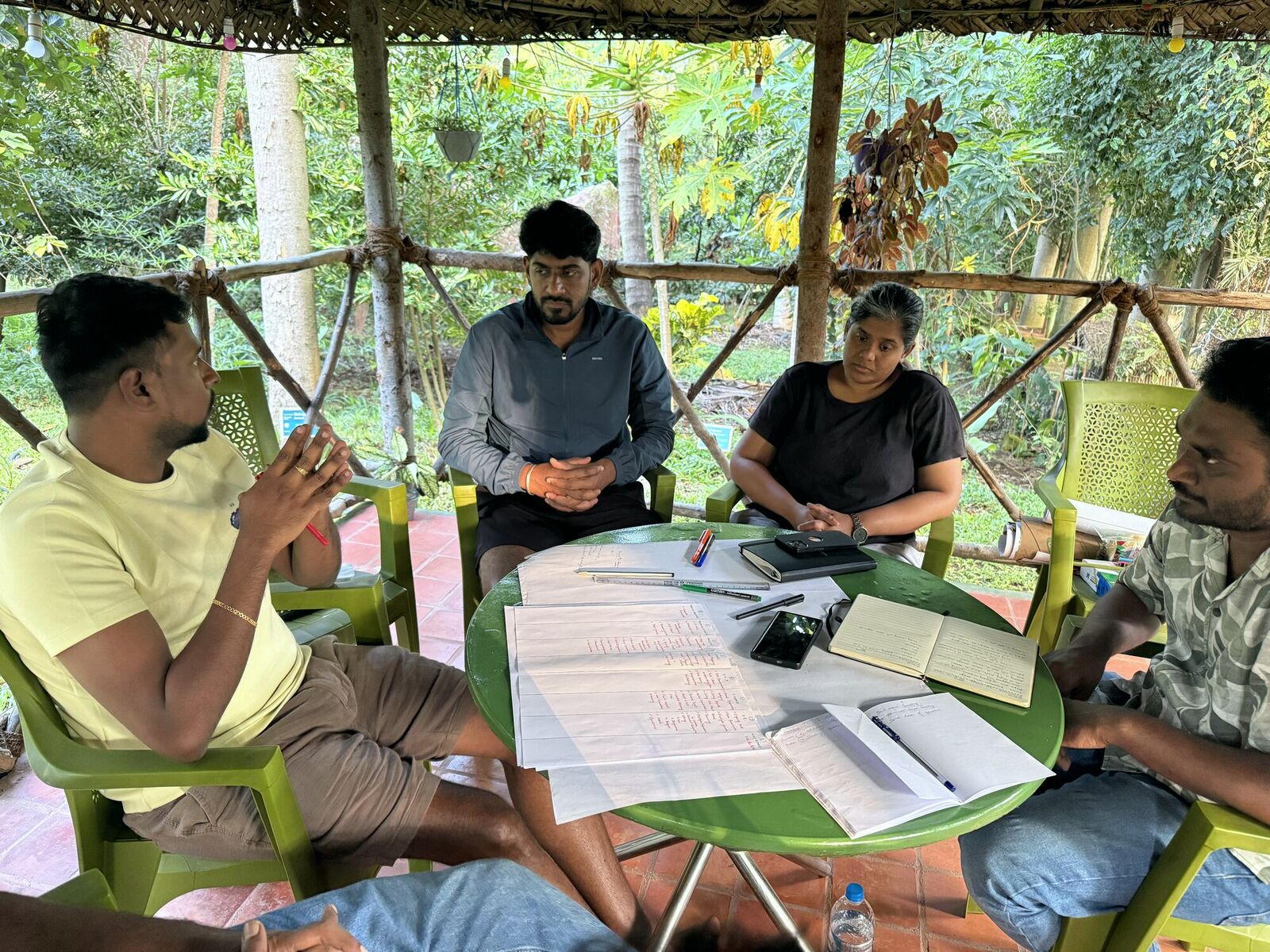 The Tribe working through plans around a table in an open forest hut